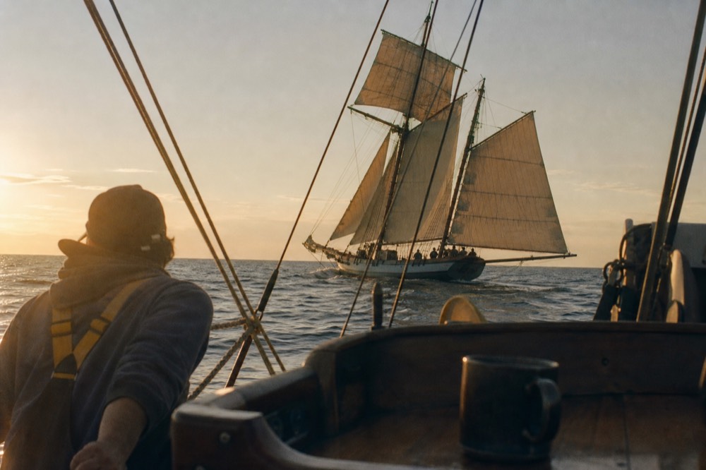 Two-masted tall ship with tan sails raised, gliding through golden evening light, no text, no logos, no people