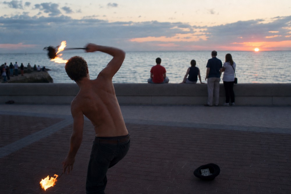 A performer juggling fire along a crowded seawall as the sun sets over the Gulf, no text, no logos, no people