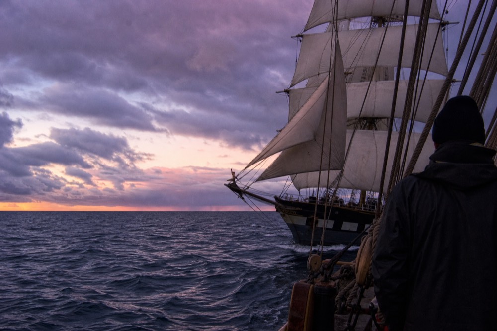 A tall sailing ship with full sails moving across open water at dusk, no text, no logos, no people