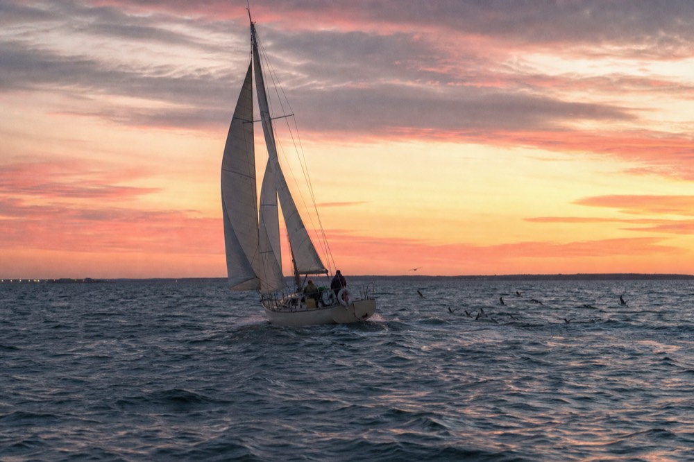 A sailboat on open water under a wide pink and orange evening sky, no text, no logos, no people